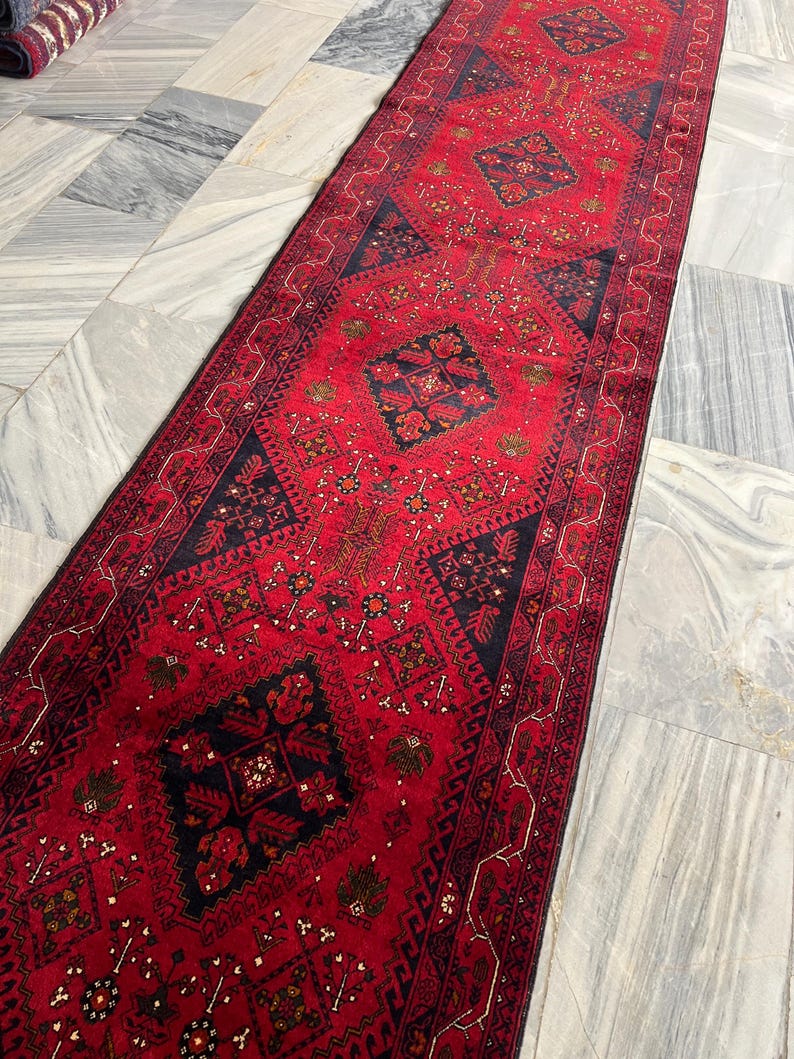 Op de afbeelding: Een lange, smalle loper met een levendige rode achtergrond en herhalende donkerblauwe geometrische ruitmotieven. Het kleed is versierd met ingewikkelde, kleinschalige bloem- en tribale patronen in tinten goud, wit en zwart.