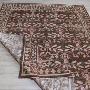 May include: A rectangular area rug featuring a deep brown background with a repeating floral and vine pattern in shades of cream and muted red. The rug has a wide decorative border and one corner is folded over to reveal the lighter underside.