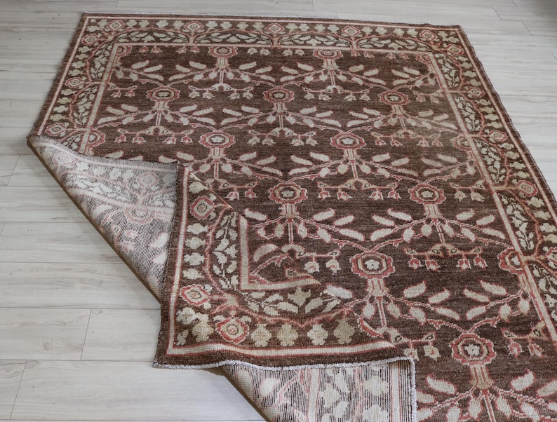 May include: A rectangular area rug featuring a deep brown background with a repeating floral and vine pattern in shades of cream and muted red. The rug has a wide decorative border and one corner is folded over to reveal the lighter underside.