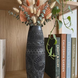 Puede incluir: Un jarrón cilíndrico alto de color negro con intrincados patrones geométricos y florales tallados. Está lleno de un arreglo floral seco que incluye colas de conejo de color beige y óxido, ramas de eucalipto y dos hojas de palma en abanico.