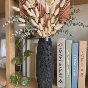 May include: A tall black cylindrical vase with an intricate carved floral pattern, filled with a dried floral arrangement of beige pampas grass, fluffy bunny tails, brown fan-shaped palm leaves, and green eucalyptus stems.