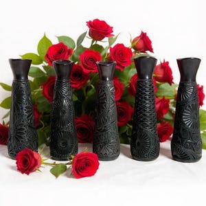 K&ouml;nnte beinhalten: F&uuml;nf schwarze Zier-Vasen mit aufwendigen gepr&auml;gten Mustern sind in einer Reihe angeordnet. Dahinter bildet eine &uuml;ppige Anordnung roter Rosen mit gr&uuml;nen Bl&auml;ttern einen lebendigen Hintergrund. Die Vasen haben eine dunkle, matte Oberfl&auml;che.