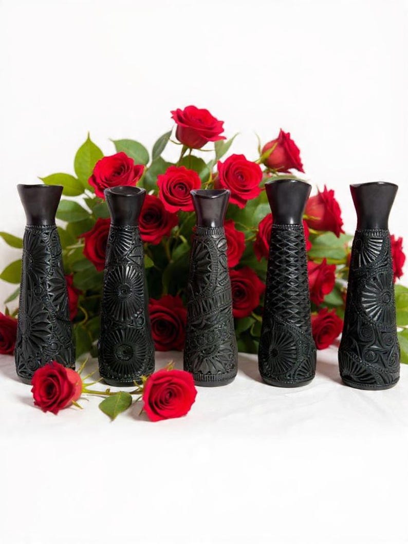 K&ouml;nnte beinhalten: F&uuml;nf schwarze Zier-Vasen mit aufwendigen gepr&auml;gten Mustern sind in einer Reihe angeordnet. Dahinter bildet eine &uuml;ppige Anordnung roter Rosen mit gr&uuml;nen Bl&auml;ttern einen lebendigen Hintergrund. Die Vasen haben eine dunkle, matte Oberfl&auml;che.