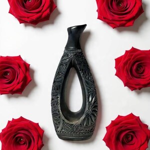 K&ouml;nnte beinhalten: Eine schwarze, geschnitzte Vase mit offenem Zentrum, umgeben von sechs leuchtend roten Rosen auf wei&szlig;em Hintergrund. Die Vase weist komplizierte Blumen- und Bl&auml;ttermuster auf, die einen auff&auml;lligen Kontrast zu den Rosen bilden. Die Vase ist etwa 25 cm hoch.