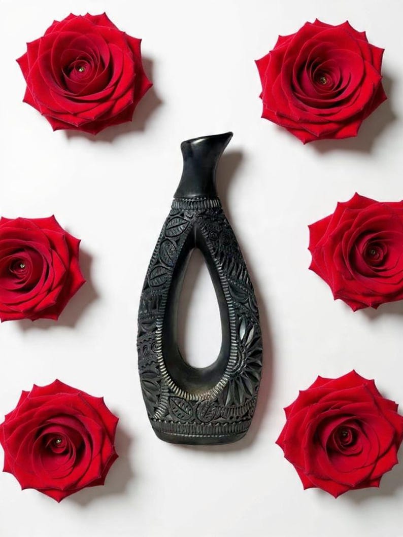 K&ouml;nnte beinhalten: Eine schwarze, geschnitzte Vase mit offenem Zentrum, umgeben von sechs leuchtend roten Rosen auf wei&szlig;em Hintergrund. Die Vase weist komplizierte Blumen- und Bl&auml;ttermuster auf, die einen auff&auml;lligen Kontrast zu den Rosen bilden. Die Vase ist etwa 25 cm hoch.