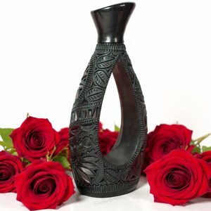 K&ouml;nnte beinhalten: Eine schwarze, geschnitzte Vase mit offenem Zentrum, die komplizierte Blumen- und geometrische Muster aufweist. Die Vase ist von leuchtend roten Rosen umgeben, die einen auff&auml;lligen Kontrast zum wei&szlig;en Hintergrund bilden. Die Vase ist etwa 25 cm hoch.