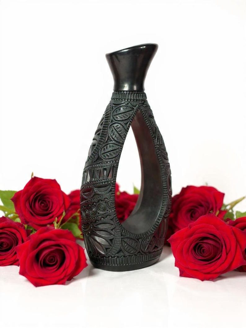K&ouml;nnte beinhalten: Eine schwarze, geschnitzte Vase mit offenem Zentrum, die komplizierte Blumen- und geometrische Muster aufweist. Die Vase ist von leuchtend roten Rosen umgeben, die einen auff&auml;lligen Kontrast zum wei&szlig;en Hintergrund bilden. Die Vase ist etwa 25 cm hoch.
