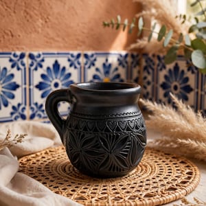 Może przedstawiać: Matowy czarny kubek ceramiczny o zaokrąglonym korpusie, ozdobiony misternym rzeźbionym wzorem geometrycznych płatków oraz pasem małych serc przy krawędzi. Kubek posiada solidny uchwyt z boku oraz gładkie, ciemne wykończenie na całej powierzchni.
