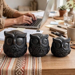 May include: A set of three dark charcoal-colored owl figurines of varying sizes, each featuring intricate carved geometric patterns on the body and circular spiral details around the eyes, arranged in a row on a flat surface.