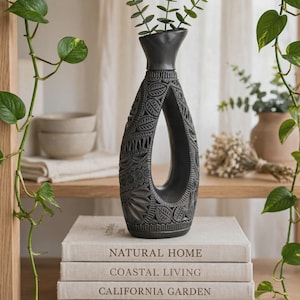 May include: A matte black ceramic vase with a hollow centre and intricate carved leaf and geometric patterns. The vase has a tapered neck and holds a small green sprig. It is positioned on a stack of four books titled Natural Home, Coastal Living, California Garden, and Craft & Clay.