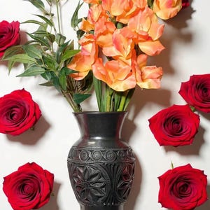 May include: A black vase with a floral design holds orange and green flowers. Red roses are scattered around the vase on a white background. The vase is approximately 8 inches tall.