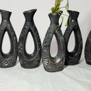 K&ouml;nnte beinhalten: Eine Sammlung dunkelgrauer Vasen mit aufwendigen, geschnitzten Blumen- und geometrischen Mustern. Jede Vase hat ein einzigartiges offenes Design und einen schmalen Hals. Eine Vase enth&auml;lt Gr&uuml;n mit roten Beeren, was dem Arrangement Farbe verleiht.