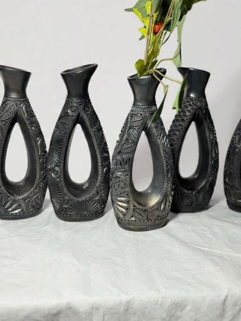 K&ouml;nnte beinhalten: Eine Sammlung dunkelgrauer Vasen mit aufwendigen, geschnitzten Blumen- und geometrischen Mustern. Jede Vase hat ein einzigartiges offenes Design und einen schmalen Hals. Eine Vase enth&auml;lt Gr&uuml;n mit roten Beeren, was dem Arrangement Farbe verleiht.