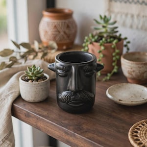 May include: A matte black ceramic planter shaped like a human head with a prominent nose, closed eyes, and a detailed mustache. The vessel features two small protruding handles on the sides and a wide circular opening at the top for plants.