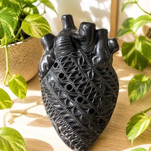 Może przedstawiać: Czarna rzeźba anatomicznego serca z misternymi rzeźbionymi wzorami na powierzchni. Obiekt ma gładkie, ciemne wykończenie z kilkoma małymi okrągłymi otworami wkomponowanymi w szczegółowy, powtarzalny geometryczny wzór.