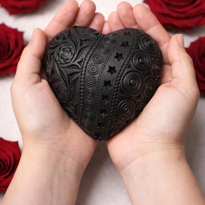 Puede incluir: Un objeto decorativo negro en forma de corazón con intrincados patrones en relieve, incluyendo espirales, círculos y estrellas. El objeto se sostiene en manos ahuecadas, con rosas rojas en el fondo. El corazón es un símbolo de amor y afecto.
