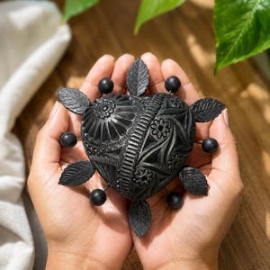 May include: A black heart-shaped ornament featuring detailed floral and geometric designs. The ornament is embellished with small black leaves and spheres. The heart is held in open hands, with a wooden surface and green foliage in the background.