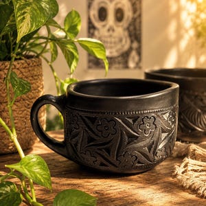 Może przedstawiać: Matowy czarny kubek ceramiczny o szerokim, niskim profilu z uchwytem po lewej stronie. Zewnętrzna powierzchnia jest ozdobiona rzeźbionym, powtarzającym się wzorem pięciopłatkowych kwiatów oraz geometrycznych trójkątnych kształtów wokół korpusu.