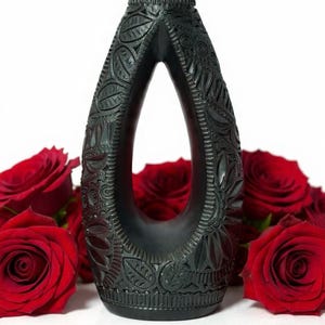 K&ouml;nnte beinhalten: Eine schwarz gef&auml;rbte, aufwendig geschnitzte Vase mit tropfenf&ouml;rmiger &Ouml;ffnung. Die Vase weist detaillierte Blumen- und Bl&auml;ttermuster auf. Rote Rosen sind um die Basis der Vase angeordnet und bilden einen starken Kontrast.