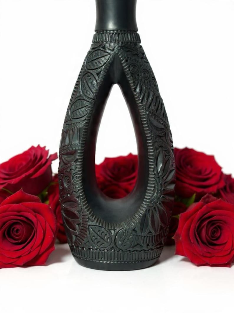 K&ouml;nnte beinhalten: Eine schwarz gef&auml;rbte, aufwendig geschnitzte Vase mit tropfenf&ouml;rmiger &Ouml;ffnung. Die Vase weist detaillierte Blumen- und Bl&auml;ttermuster auf. Rote Rosen sind um die Basis der Vase angeordnet und bilden einen starken Kontrast.