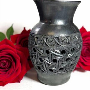 Może przedstawiać: Ciemnoszary ceramiczny wazon z misternymi, rzeźbionymi geometrycznymi wzorami. Wazon ma szeroką podstawę, rozszerzającą się szyję i ozdobny pas z okręg&oacute;w. Czerwone r&oacute;że w tle.