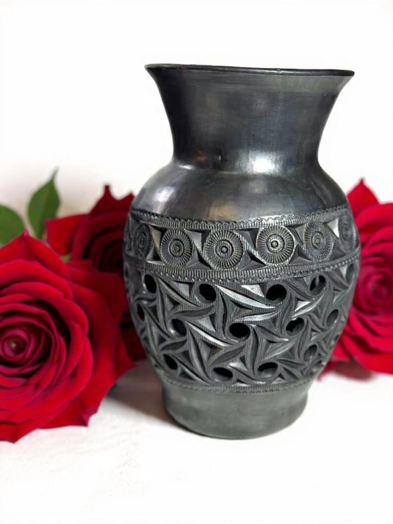 Może przedstawiać: Ciemnoszary ceramiczny wazon z misternymi, rzeźbionymi geometrycznymi wzorami. Wazon ma szeroką podstawę, rozszerzającą się szyję i ozdobny pas z okręg&oacute;w. Czerwone r&oacute;że w tle.