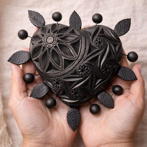 May include: A black heart-shaped decorative object with intricate floral and geometric patterns. The object is adorned with black leaves and spherical accents. The heart is held in open hands, showcasing its detailed design and texture.