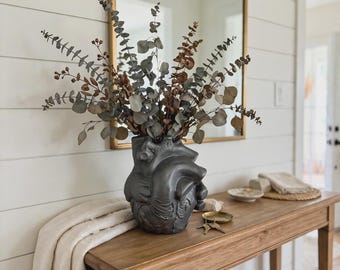 May include: A dark gray anatomical heart-shaped vase holding a bouquet of dried eucalyptus branches with green and brown leaves. The vase features realistic sculpted details of heart chambers and vessels, providing a textured, matte finish.