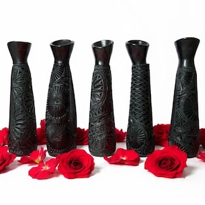 May include: Five black decorative vases with intricate carved designs, each with a unique pattern. Red rose petals and whole roses are scattered around the vases on a white background. The vases have a tapered shape.