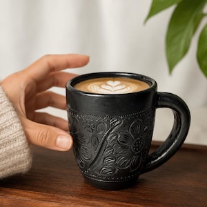Peut inclure: Tasse à café en céramique noire avec un motif floral en relief, remplie de café et de latte art. La tasse est posée sur un plateau en bois. La poignée est sur le côté droit.