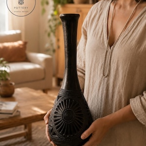 Könnte beinhalten: Eine hohe, schwarze Vase mit engem Hals, die mit komplizierten, geschnitzten kreisförmigen Blumenmustern und geometrischen Details an der Basis verziert ist. Die Oberfläche ist matt und das Design enthält ein Logo mit XQUENDA POTTERY OAXACA, MEXICO.