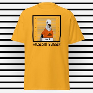 May include: Mustard yellow t-shirt featuring a graphic of a seagull in an orange shirt, framed as a mugshot. The seagull has a brown stain on its shirt. The text "No. 2" and "WHOSE $#!T IS BIGGER?" are below the image.