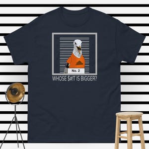 May include: Navy blue t-shirt featuring a cartoon seagull in an orange shirt with a poop graphic. The seagull is framed with the text "No. 2" and "WHOSE SH#T IS BIGGER?" printed below. The shirt is a heavy cotton blend.