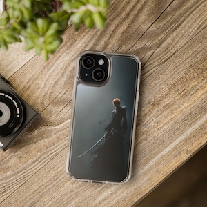 Op de afbeelding: Transparante telefoonhoes met een donkere illustratie van een personage met oranje haar en een zwaard. De hoes ligt op een houten oppervlak met een camera en groen in de achtergrond.