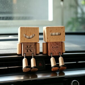 May include: Two wooden car vent ornaments shaped like robots. One has the name "Kearney" and the letter "K" engraved on its chest. The other has the initials "E&D" inside a heart. Both have smiling faces and dangling legs.