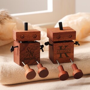 May include: Two wooden robot figurines with personalized engravings. The robots are made of dark brown wood, with black accents for the arms and a small antenna. One robot has "S/N Shelton" engraved, the other has "Kearney". The robots have wooden legs.
