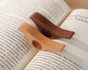 Puede incluir: Dos sujetapáginas de madera, uno claro y otro oscuro, descansan sobre un libro abierto. El sujetapáginas oscuro tiene grabado el texto "One More Page...". Los sujetapáginas están diseñados para mantener un libro abierto.