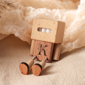 May include: A wooden robot-shaped toy with a light brown head and body, featuring a rectangular head with a mouth-like slot and white teeth. The body has a letter "K" and a leaf design. The legs are made of wood and metal.