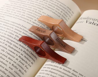 Könnte beinhalten: Drei hölzerne Buchseitenhalter in verschiedenen Braun- und Rottönen liegen auf einem offenen Buch. Einer ist mit "Jacob" graviert, ein anderer mit "Brown". Die Halter haben ein Fingerloch.
