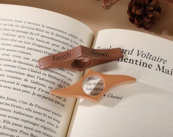 Könnte beinhalten: Zwei hölzerne Buchseitenhalter. Einer ist dunkelbraun mit dem eingravierten Namen "Jacob". Der andere ist hellbraun. Beide haben eine kreisförmige Öffnung für den Finger und liegen auf einem offenen Buch.