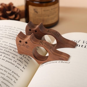 Puede incluir: Dos sujetapáginas de libros de madera con forma de gato con los nombres grabados "Kendal" y "Selina". Los sujetapáginas están sobre un libro abierto con texto impreso. La madera tiene un tono marrón cálido.