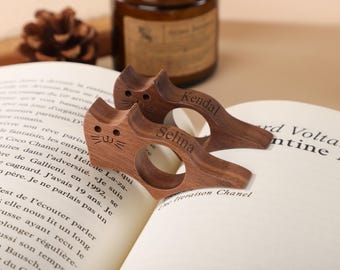 Puede incluir: Dos sujetapáginas de libros de madera con forma de gato con los nombres grabados "Kendal" y "Selina". Los sujetapáginas están sobre un libro abierto con texto impreso. La madera tiene un tono marrón cálido.