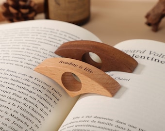 May include: Two wooden thumb page holders rest on an open book. One is light-colored with "Reading is life" engraved. The other is a darker wood. The book's pages are filled with text.