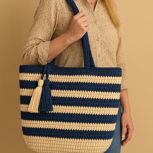 Puede incluir: Un bolso tote de crochet a rayas azul marino y crema, con asas y borlas azul marino. El bolso es de gran tamaño, ideal para llevar objetos. El bolso está hecho de un material suave y texturizado.