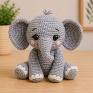 May include: A handmade gray crochet elephant with large ears, a trunk, and big black eyes. The elephant has pink cheeks and white accents on its feet. It is sitting on a wooden surface.