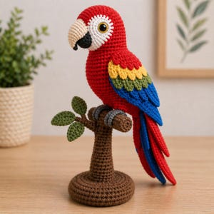 Könnte beinhalten: Ein handgehäkelter Papagei in Rot, Gelb, Blau und Grün, der auf einem gehäkelten braunen Ast sitzt. Der Vogel hat einen beigen Schnabel und Augendetails. Der Papagei ist etwa 25 cm hoch und sitzt auf einem gehäkelten braunen Sockel.