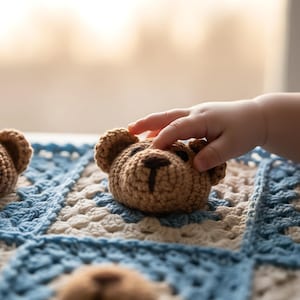 Puede incluir: Cabezas de osos de peluche marrones de ganchillo sobre una manta de ganchillo azul, blanca y beige. Las cabezas de los osos tienen ojos negros y una nariz cosida. Una mano se extiende para tocar una de las cabezas de los osos.