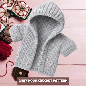 Könnte beinhalten: Ein hellgrauer Baby-Hoodie mit kurzen Ärmeln aus grobem Strickgarn. Der Hoodie hat eine Kapuze und Knöpfe vorne. Der Text "BABY HOOD CROCHET PATTERN" befindet sich am unteren Rand des Bildes. Häkelgarn und ein Häkelprojekt befinden sich im Hintergrund.