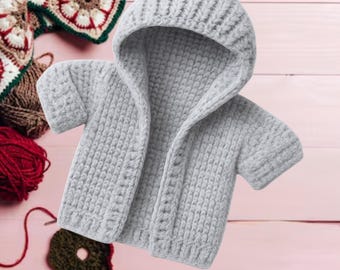 May include: A light gray, short-sleeved baby hoodie made of chunky knit yarn. The hoodie has a hood and buttons down the front. The text "BABY HOOD CROCHET PATTERN" is at the bottom of the image. Crochet yarn and a crochet project are in the background.