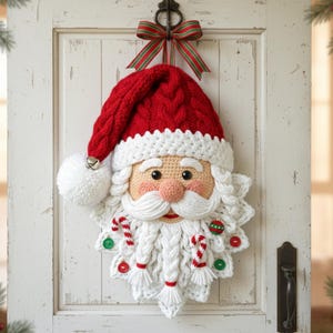 Puede incluir: Una corona navideña hecha a mano con la cara de Papá Noel. La corona presenta un gorro rojo tejido con un ribete blanco, una barba blanca y una cara sonrosada. Bastones de caramelo y adornos están unidos a la barba.